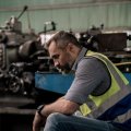 A bearded man in a short and high vis vest sits with his head bowed in a factory.